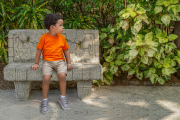 A concerned little boy sets on a cement park bench looking away.