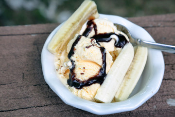 Hard ice cream just starting to melt, drizzled with chocolate sauce and garnished with banan slices in a white bowl on a painted brown wood surface with wear