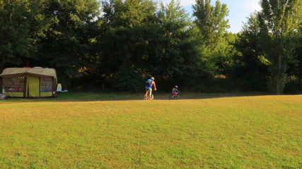 Paisaje verde con familia en bicicleta y caravana en el fondo y arboles y cielo azul al atardecer en verano de vacaciones