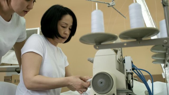 Professional asian seamstress using overlock sewing machine with assistance of female colleague in textile factory