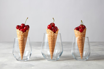 Three waffle cones with fresh ripe cherries in glasses on a gray marble table.