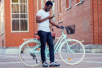 handsome African American man stylish well-dressed standing with a blue bicycle and holding phone...