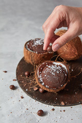 A woman decorates a homemade chocolate ice cream in a coconut with chocolate balls on a board on a gray stone background.
