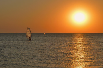 the silhouette of a windsurfer on a board under a sail moves along a calm water surface at sunset...