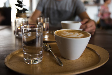 Cup of coffee cappuccino and a glass of water on a wooden tray. A man holding serving cup of coffee on the background