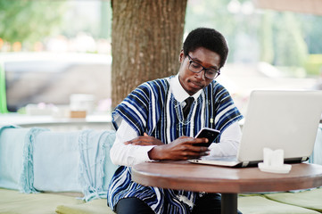 African man in traditional clothes and glasses sitting behind laptop at outdoor caffe and looking...