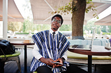 African man in traditional clothes and glasses sitting behind laptop at outdoor caffe with  mobile...
