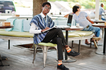 African man in traditional clothes and glasses sitting behind laptop at outdoor caffe and writing...