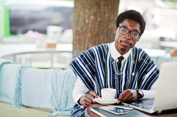 African man in traditional clothes and glasses sitting at outdoor caffe, drink coffee and working...