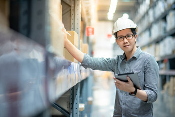 Young Asian man worker wearing safety helmet and eyeglasses doing stocktaking of product in cardboard box on shelves in warehouse by using digital tablet and pen. Physical inventory count concept