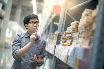 Young Asian man worker doing stocktaking of product in cardboard box on shelves in warehouse by...