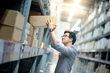 Young Asian man taking cardboard box from shelves in warehouse. Shopping warehousing or working...