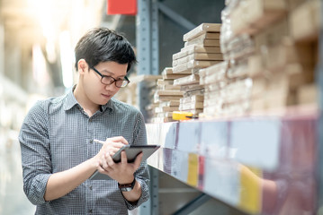 Young Asian man worker doing stocktaking of product in cardboard box on shelves in warehouse by using digital tablet and pen. Physical inventory count concept