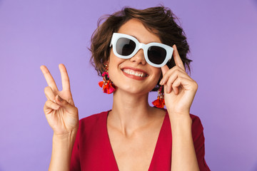 Image of fashionable brunette woman 20s in straw hat and sunglasses showing peace sign, isolated over violet background