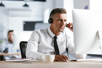 Photo of caucasian man 30s wearing office clothes and headset, sitting by computer in call center