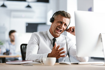 Photo of nervous businessman 30s wearing office clothes and headset, yelling while sitting by computer