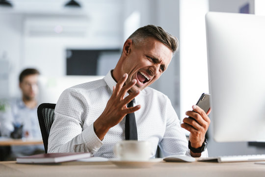 Image of annoyed businessman 30s wearing white shirt and tie sitting at desk in office by computer, and yelling while talking on smartphone
