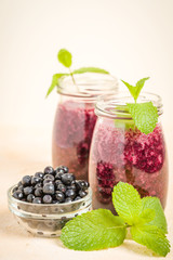 Blueberry smoothie decorated with fresh green mint leaves and raw ripe berries on yellow pastel background.
