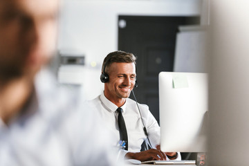 Photo of businesslike man 30s wearing office clothes and headset, smiling while sitting by computer in call center