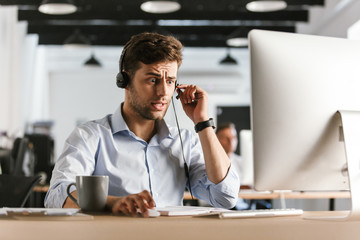 Photo of stressed man 30s wearing office clothes and headset, sitting by computer and working in...