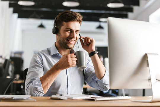 Photo of businesslike man 20s wearing office clothes and headset, drinking tea from cup while sitting by computer in call center