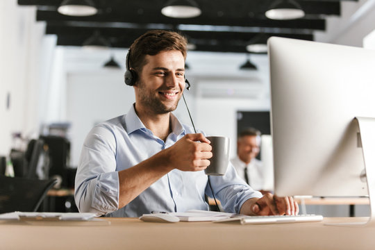 Photo of businessman 30s wearing office clothes and headset, drinking tea from cup while sitting by computer in call center