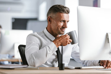 Naklejka premium Image of happy businessman 30s wearing white shirt and tie drinking tea from cup, while sitting by computer in office