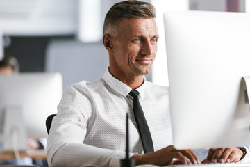 Image of european businesslike man 30s wearing white shirt and tie sitting at desk in office, and working at computer