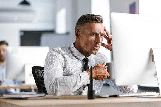 Photo of happy director man 30s wearing white shirt and tie drinking tea from cup, while sitting by computer in office