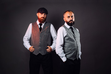 Two bearded men in strict suits standing one near one and looking on camera on black