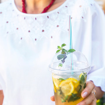 A woman in a white blouse holds a plastic glass with a mojito mug and a mint. Summer drink to cool outside