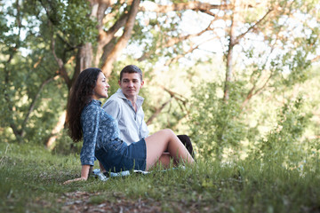 young couple sitting on the grass in the forest and looking on sunset, summer nature, bright sunlight, shadows and green leaves, romantic feelings