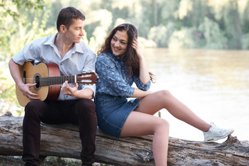 young couple sitting on a log by the river and playing guitar, summer nature, bright sunlight, shadows and green leaves, romantic feelings