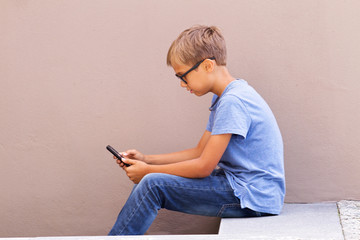 Boy with smartphone sitting on stairs outdoor. Kid wathing to screen, reading, typing, playing games