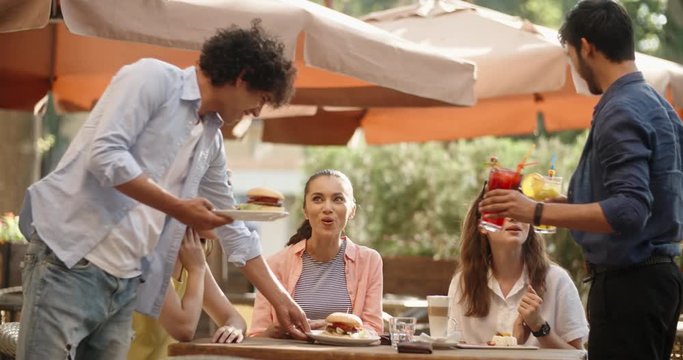 Three girls sitting at table in cafe chatting, then two guys approach with food and drinks. Friends enjoying their time 4k