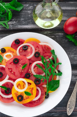 Salad of red and yellow tomatoes with olives, onions and basil. In a white big plate on a wooden background.