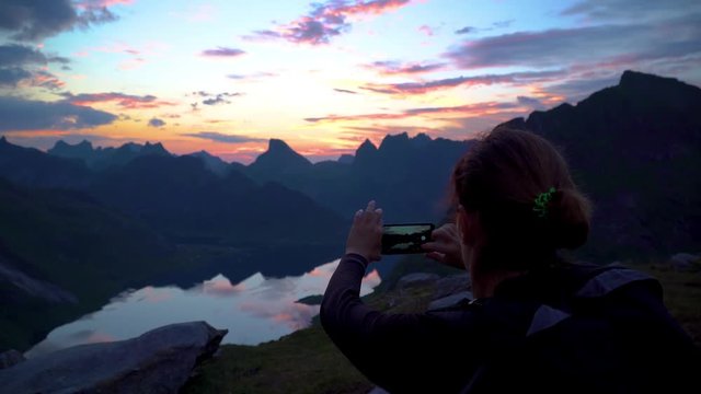 Young woman traveler on a background of mountains takes photos on a mobile phone. Dawn in the mountains and the beautiful nature of the Lofoten islands. Norway. 4K