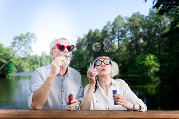 Red sunglasses. Funny elderly man wearing red heart shape sunglasses using soap bubbles standing...
