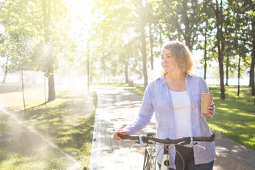 Naklejka na ściany i meble Young blonge woman walking her bicycle in the park and holding a cup of coffee