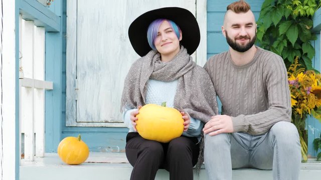 Portrait of young woman and a man, a family smiling beautiful woman with blue hair sitting on porch of country blue house, green loach plant and vase with sunflowers, yellow pumpkins. A sweater, scarf