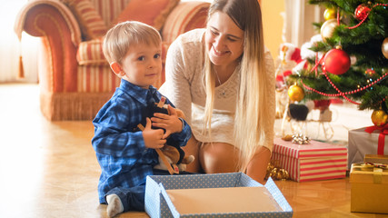 Adorable little boy sitting under Christmas tree and hugging plush toy he received in gift box from Santa