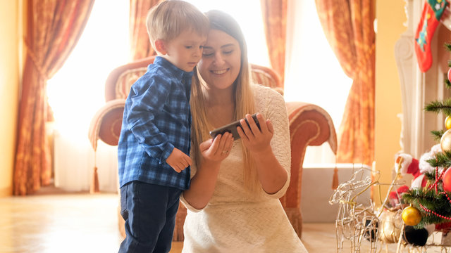 Portrait of happy young mother watching video on smartphone with her little son next to Christmas tree