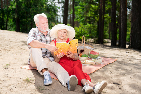 Book and beach. Couple of good-looking beaming stylish pensioners reading book while sitting on the beach