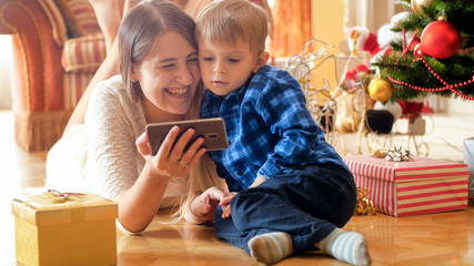 Laughing young family with little son lying on floor on Christmas morning and watching video on smart phone