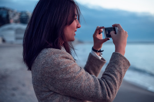 Young woman standing on beach, taking photograph of view, using smartphone