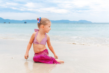 Little beautiful girl dressed in swimsuit as a mermaid sits on the seashore