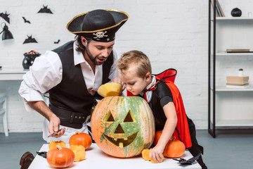 Fototapeta premium man in pirate costume and son in vampire halloween costume looking into pumpkin together at home