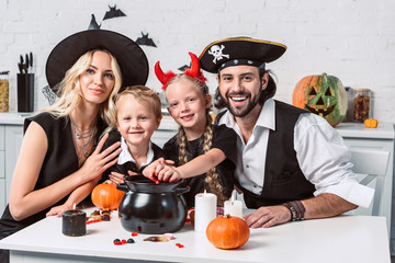 family in various halloween costumes at table with black pot with treats in kitchen at home