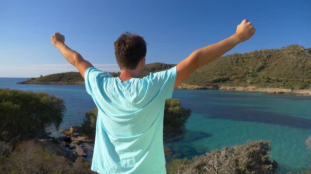 SLOW MOTION CLOSE UP: Ecstatic male tourist celebrates reaching a scenic viewpoint in spectacular Sardinia. Unrecognizable young man outstretches his arms while observing the picturesque Italian beach