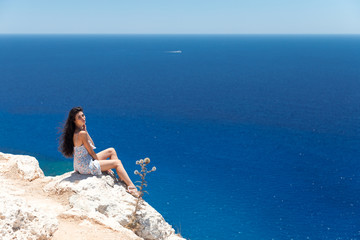Brunette in a dress sits on the edge of the cliff overlooking the sea